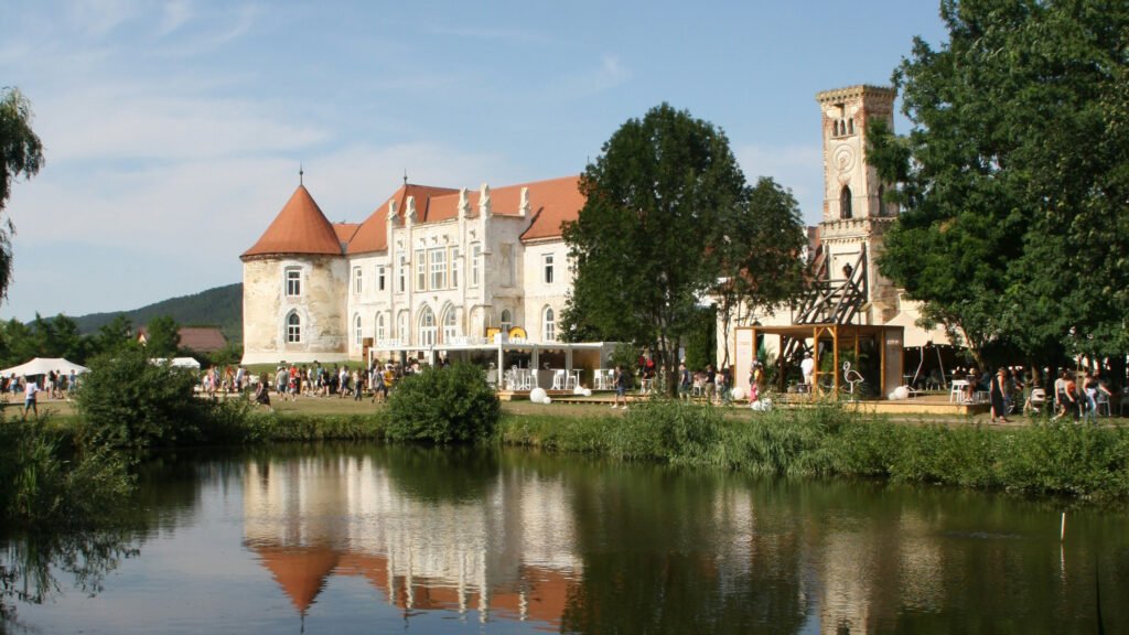 Historic Baroque-style castle with red-tiled roof, arched windows, and a reflection pond in Bontida, Romania, surrounded by trees and festival tents.