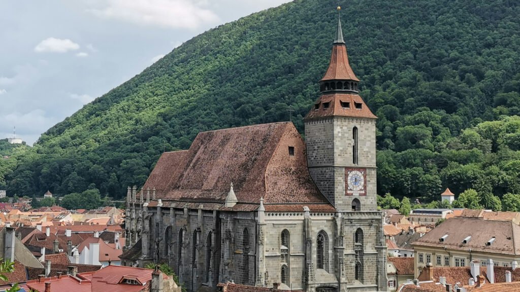 Gothic-style Black Church with a tall clock tower and steep red-tiled roof, located in Brasov, Romania, surrounded by forested hills.