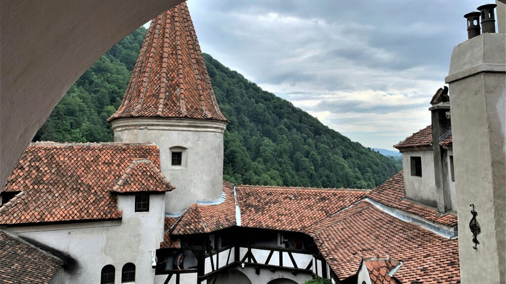 Steep red-tiled rooftops and towers of Bran Castle, surrounded by forested hills, in Bran, Romania, under a cloudy sky