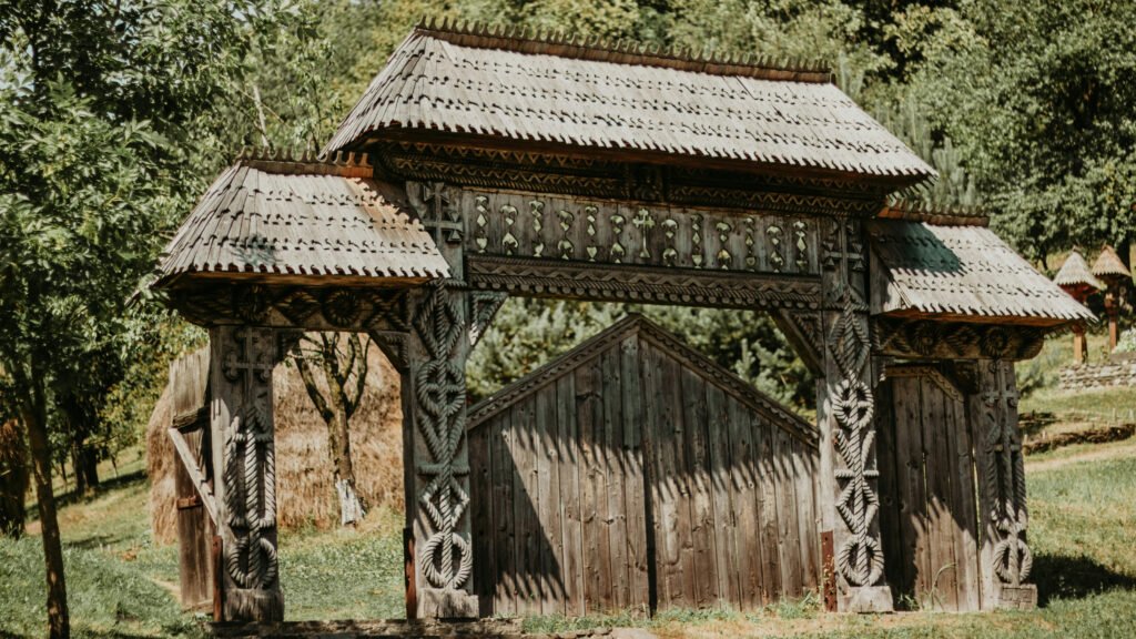 Traditional carved wooden gate with intricate geometric and rope motifs, located in Bucovina, Romania, surrounded by trees and grass.