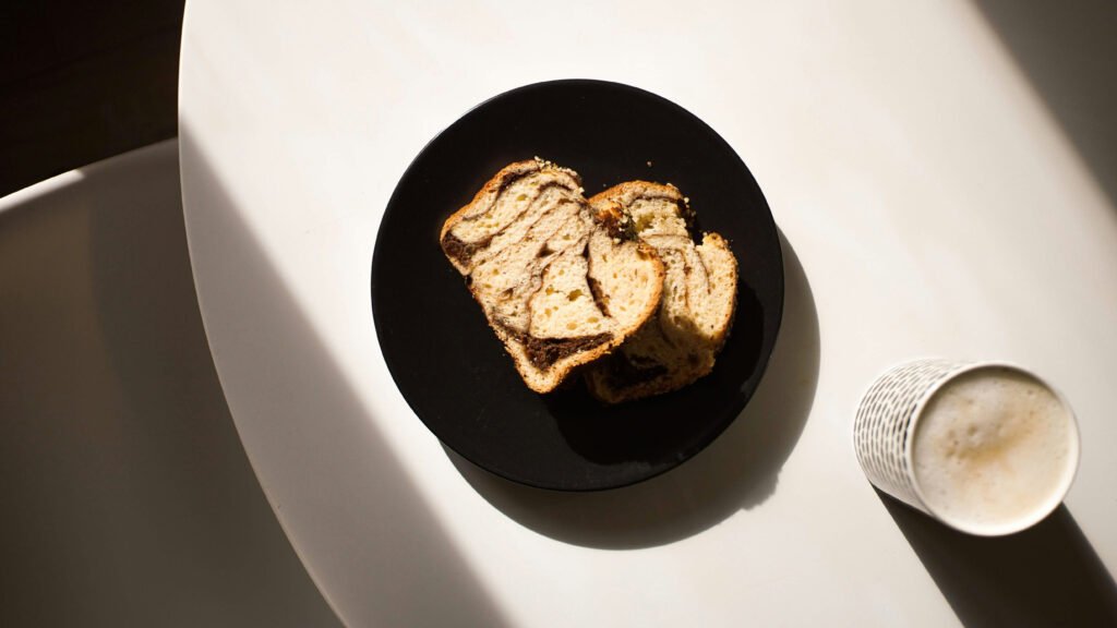 Top-down view of Romanian cozonac slices on a black plate, next to a cup of frothy coffee on a sunlit white table.