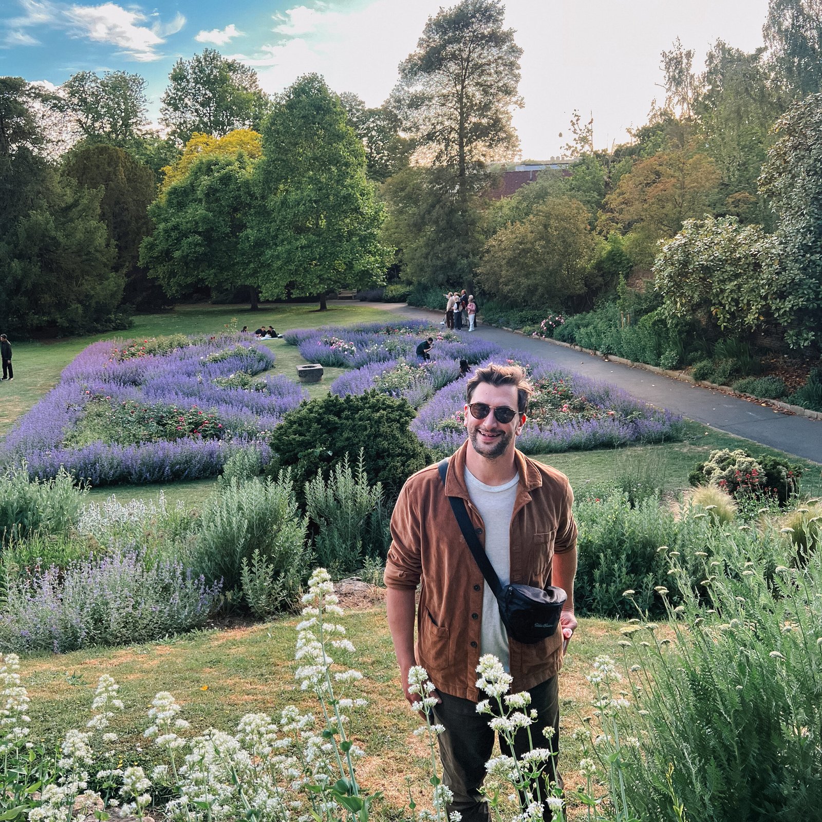 Gabriel McNab standing in front of a blooming purple flowerbed surrounded by lush greenery in a park.