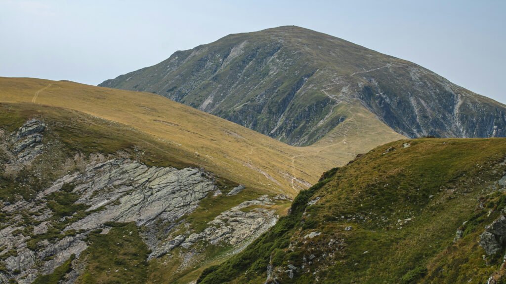 Grassy mountain ridgeline with rocky outcrops and steep slopes in the Parang Mountains, Romania, under a clear pale blue sky.