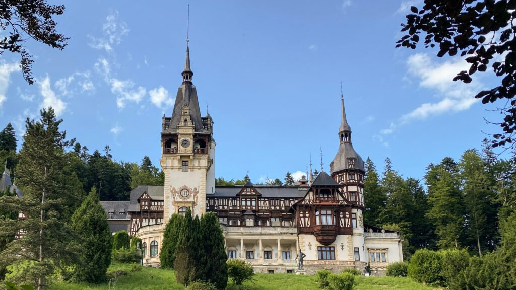 Neo-Renaissance castle with timbered facades, pointed towers, and stone base surrounded by trees in Sinaia, Romania.