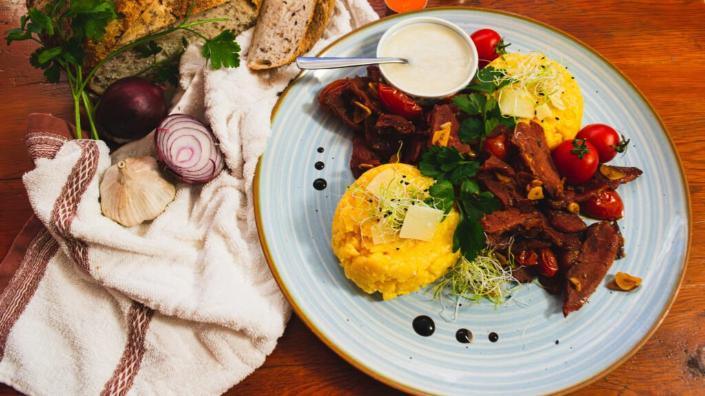 Traditional Romanian dish with polenta, slices of cured meat, cherry tomatoes, garlic, and fresh herbs on a blue ceramic plate.