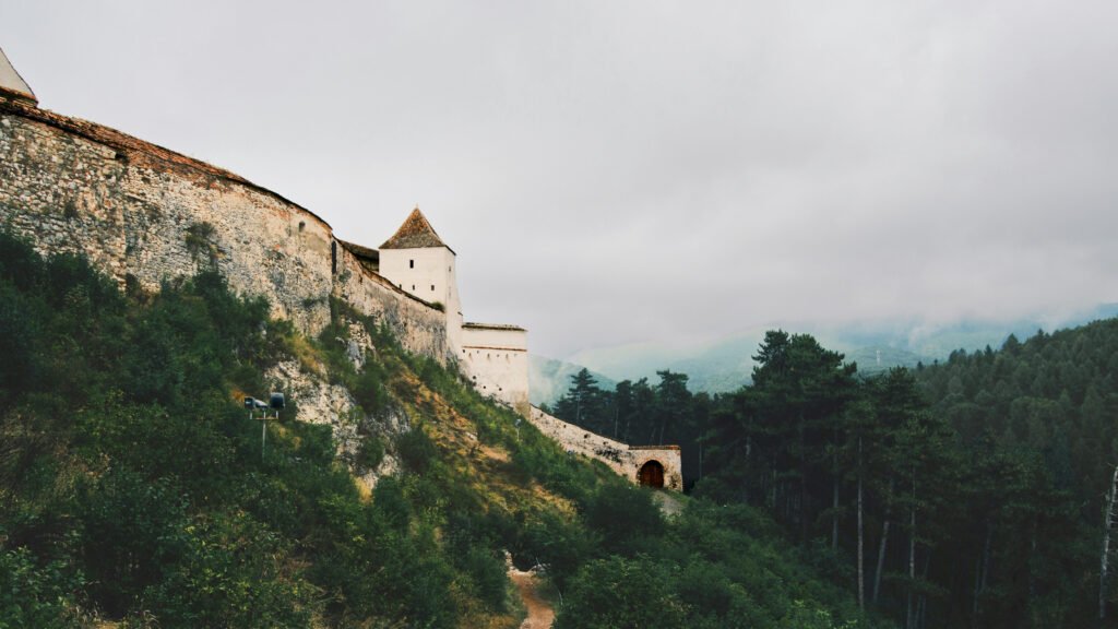 Stone fortress wall with corner tower and arched gate, built on a forested hillside in Rasnov, Romania, surrounded by misty mountain landscape.
