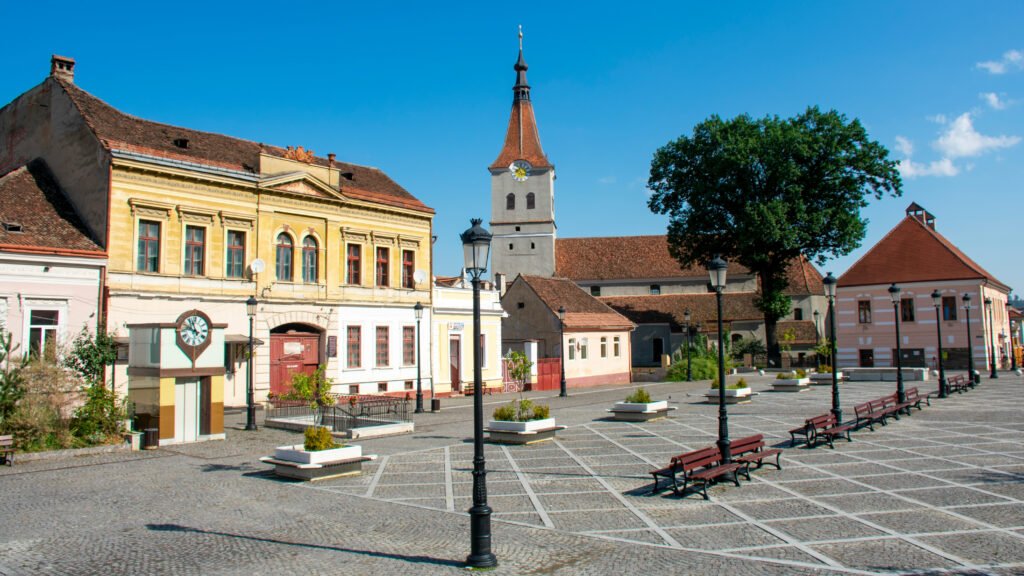 Cobblestone town square with benches, colorful historic buildings, and a church tower under a clear blue sky in Rasnov, Romania.