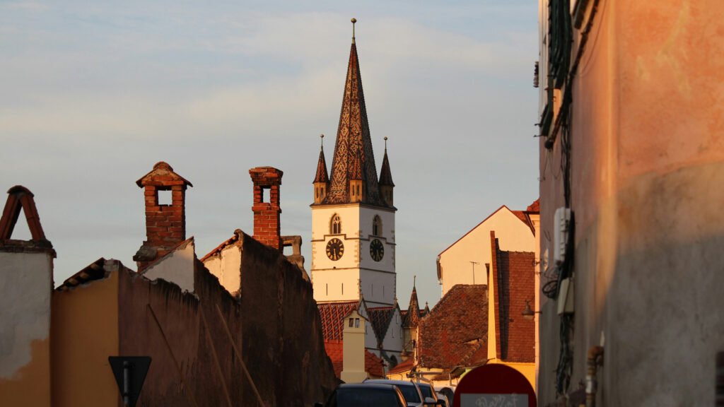Gothic-style Lutheran church tower with colorful patterned roof tiles and clock faces, rising above rooftops in Sibiu, Romania.