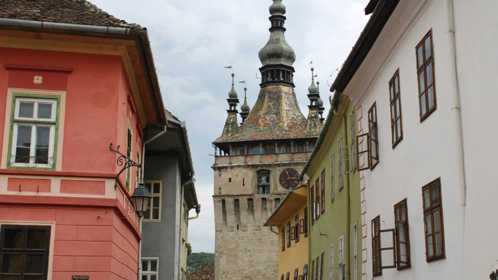 Medieval clock tower with a colorful tiled roof and spires rising above pastel buildings in Sighișoara, Romania.