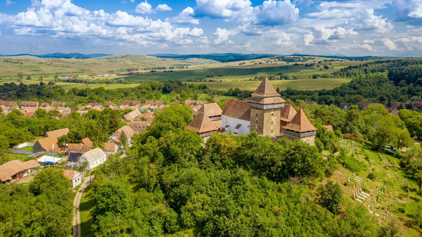 Hilltop fortified church with defensive towers and terracotta roof tiles in Viscri, Romania, surrounded by trees and rolling countryside.